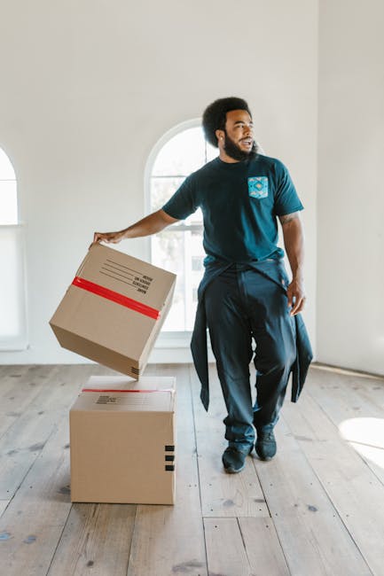 A man with a dark beard and curly hair, dressed in a navy blue t-shirt and trousers, is inside a bright, empty room with white walls and wooden flooring. He is holding a cardboard box with red tape along its sides, preparing for a home relocation or furniture transport process. Two additional cardboard boxes, also taped and of different sizes, are placed on the floor nearby. Behind him, an arched window allows natural light to enter, illuminating the scene. The presence of packing materials and the man's stance suggest he is engaged in packing or unpacking as part of a professional house removals service, with the setting indicating an indoor environment ready for a move. This visual relates to the services offered by Movers Catford, providing efficient home moving and packing solutions in the Catford SE6 area, including same-day quotes and furniture transport support.