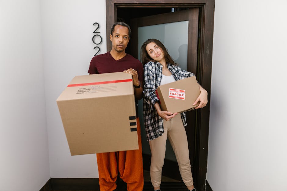 A man and a woman standing inside a residential building near a doorway during a home relocation process. The man, dressed in casual orange trousers and a maroon shirt, is holding a large cardboard box with black tape on the edges, while the woman, wearing beige trousers and a checked shirt, is holding a smaller cardboard box with 'Fragile' and red tape labels. Both are positioned on a small landing or entrance area, with a dark wooden door frame behind them. The lighting is natural, illuminating the indoor space. This image captures the loading process typical of house removals, with cardboard boxes prepared for transport, supported by the professional moving services of Movers Catford. The environment suggests careful handling and organized packing, essential elements in furniture transport and packing and moving activities related to affordable home removals in the Catford SE6 area.