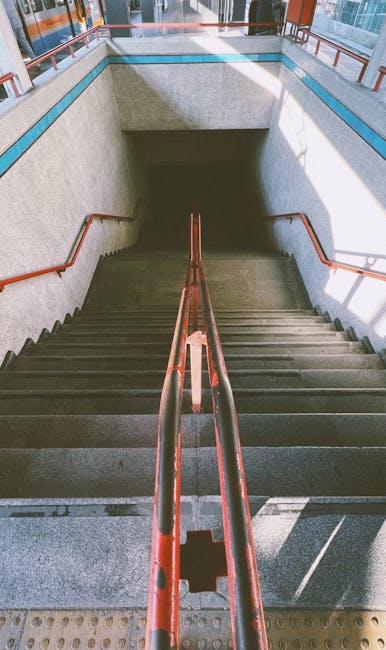 A downward view of a concrete staircase leading to an underground subway or train station, with metal handrails painted in red along both sides. The staircase is enclosed by light grey concrete walls, and at the bottom, a darkened platform or area is partially visible. Natural light illuminates the top of the stairs through openings or windows, creating a contrast between the well-lit upper section and the shadowed lower section. This scene is part of a transportation hub, potentially near Catford Bridge station, with the stairwell designed to facilitate passenger movement during a home or office relocation process. Movers Catford may use such staircases to assist with furniture transport and packing during house removals in areas with narrow access points, emphasizing the importance of careful handling and logistical planning in moving services.