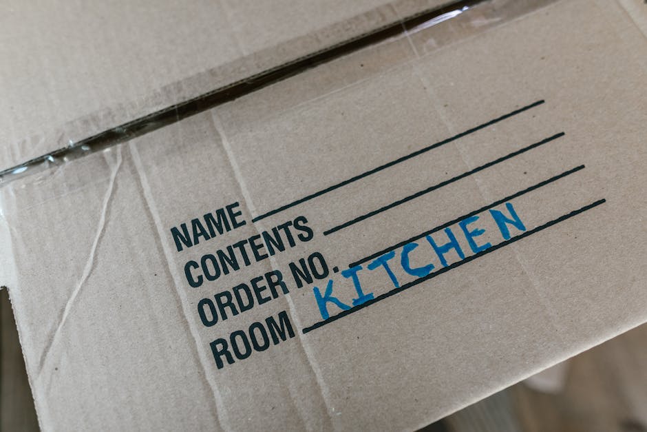 A cardboard moving box labeled with black text showing fields for 'Name', 'Contents', 'Order No.', and 'Room,' with ‘Kitchen’ handwritten in blue ink under the 'Room' label. The box is partially wrapped in clear plastic tape and is situated on a wooden floor inside a home, likely part of a furniture transport or packing process during a home relocation. The image captures the box from an angled top-down perspective, highlighting the labeling and packing materials used by Movers Catford as part of their removals services, which include careful packing and loading in preparation for a house move in Lewisham or Catford.